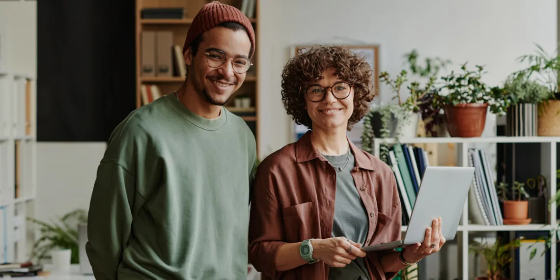 Dois profissionais sorriem em um escritório com plantas; um deles segura um notebook